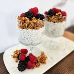 2 glass cups filled with chia pudding, topped with granola and fruit on a white marble tray